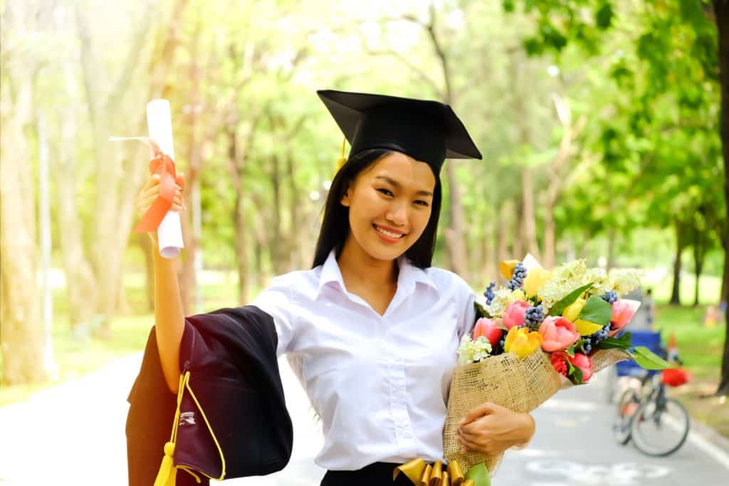 A happy graduate stands smiling with her diploma and graduation cap.