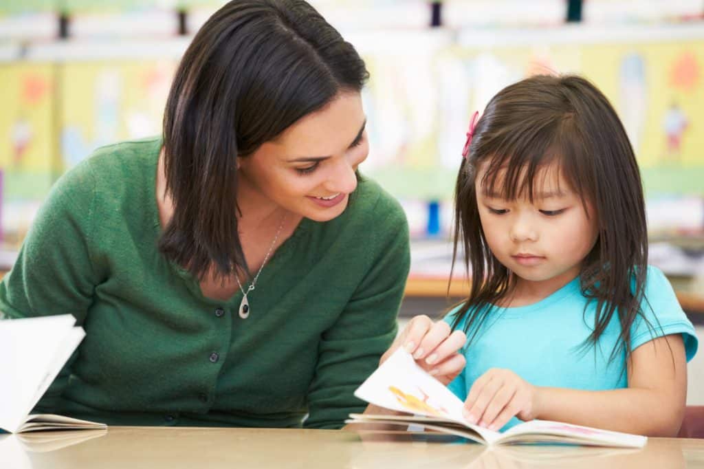 Young teacher sitting at a table with a very young student helping her read a book.