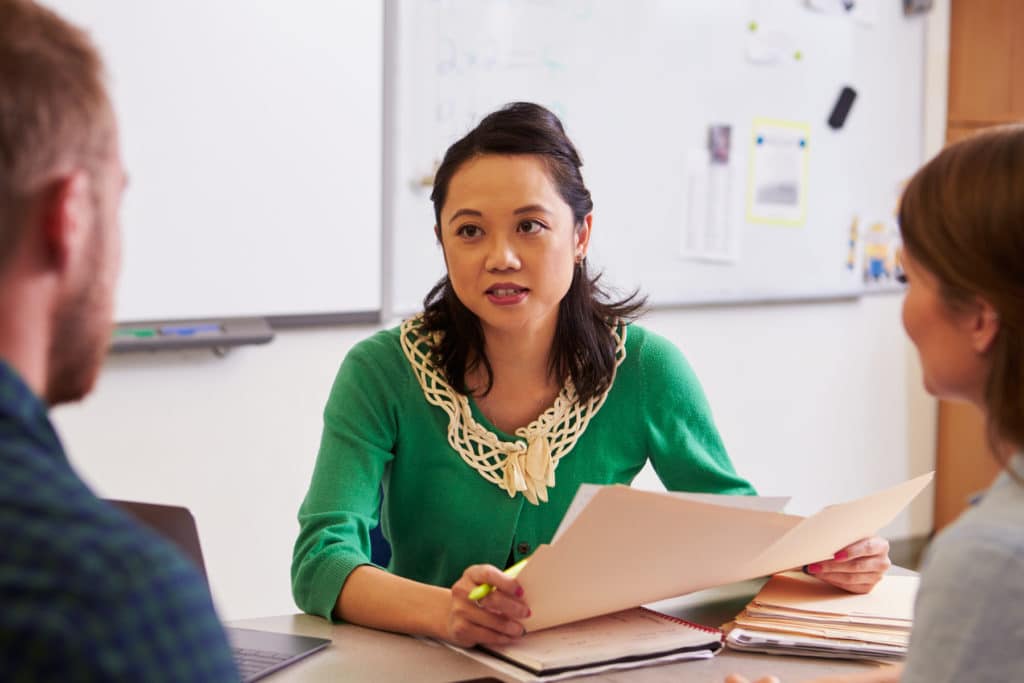 Women sitting at a desk in a classroom holding paperwork and talking to other adults.