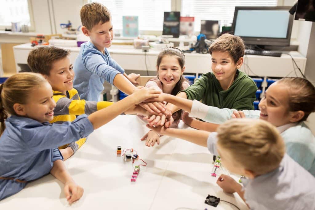 A group of happy students do a hand shake over the robot they are building in the classroom.