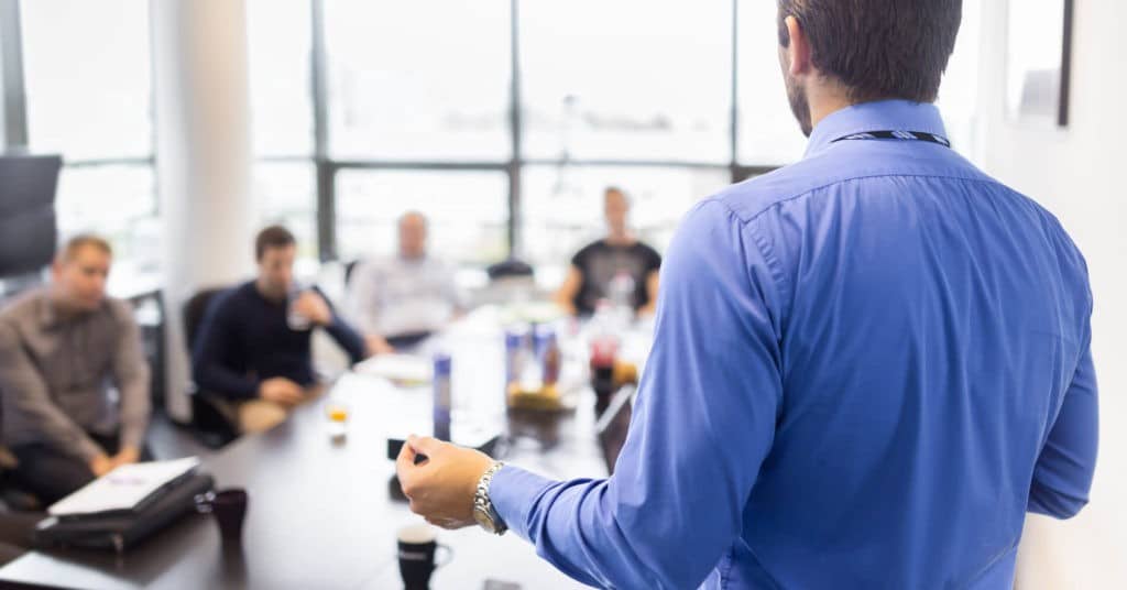 Man talking and standing in front of other people seated at a table.