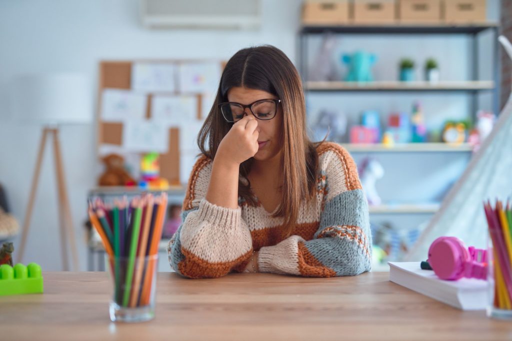 A teacher sits at a school desk, holding her nose, appearing stressed.