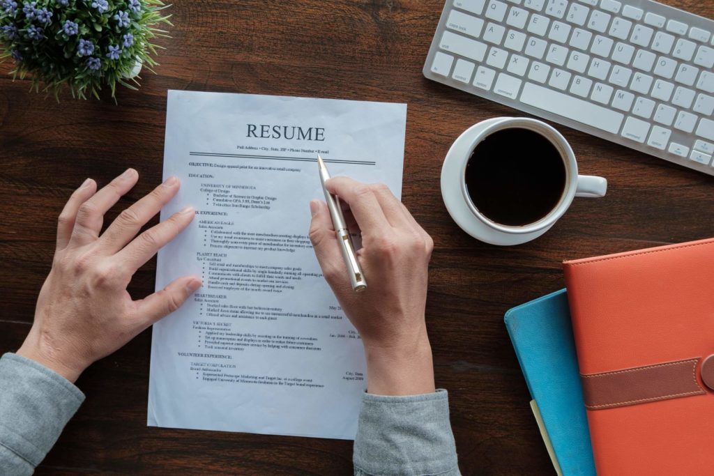 Someone holds a resume application with a pen; a coffee cup, keyboard, and notebook are in the background.
