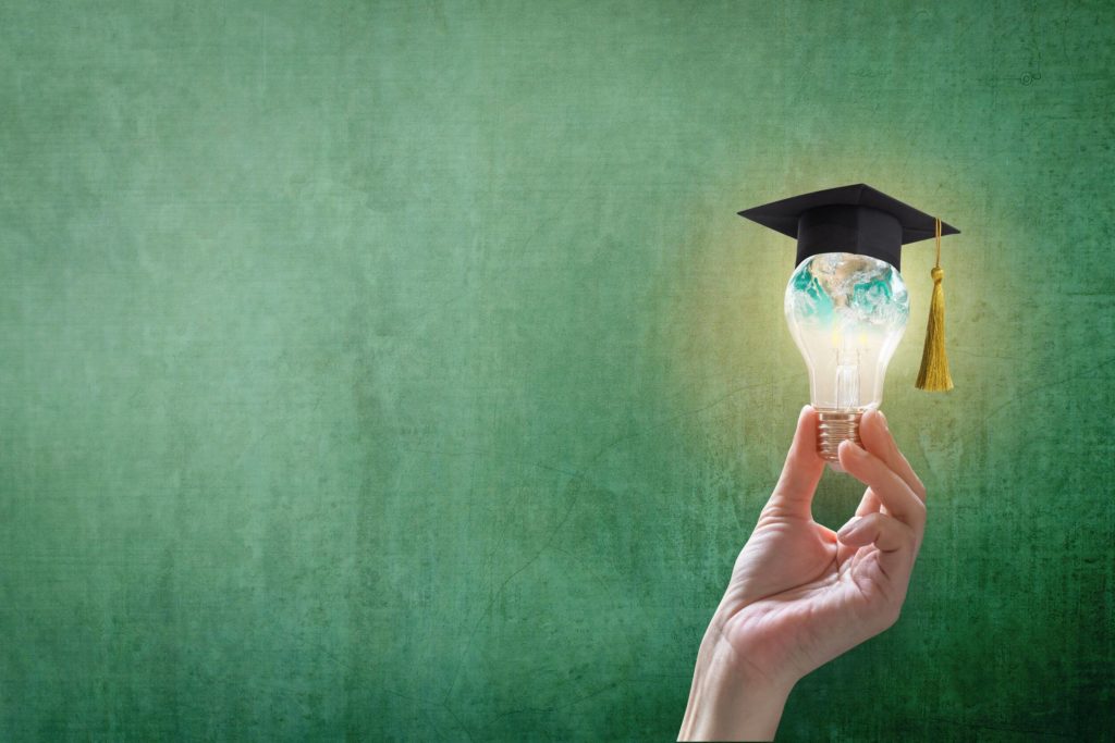 A hand holds a light build with a graduation cap on top in front of a teacher chalkboard.
