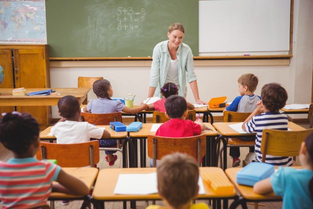 Pupils listen to their teacher talking by the chalkboard at the front of the classroom.