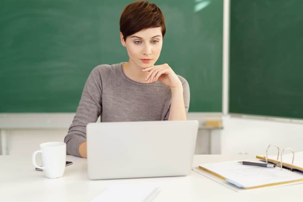 Female teacher sitting at a desk in a classroom looking at a laptop.