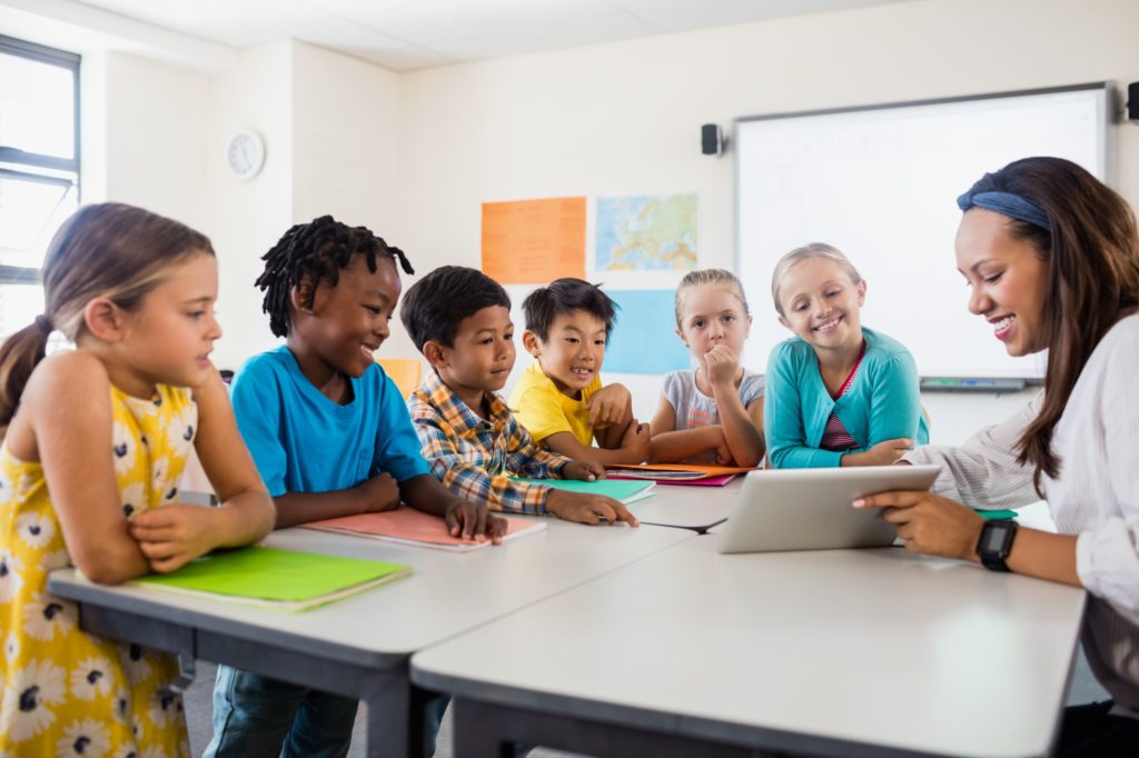 Young students sitting at a table in a classroom with a teacher showing them something on a tablet.