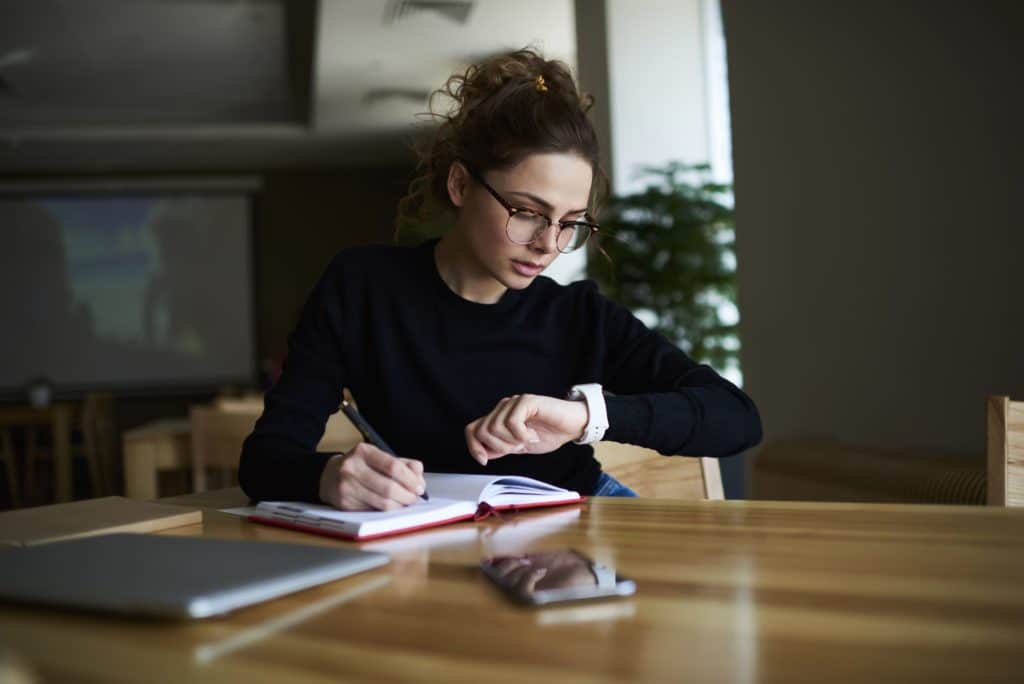 A female student looks at her watch while writing in a notebook.