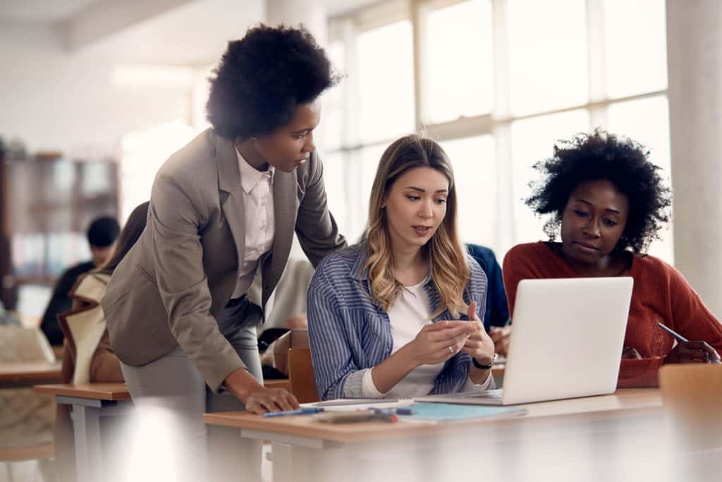 Women students ask their teacher a question, who is leaning over to look at their computer.