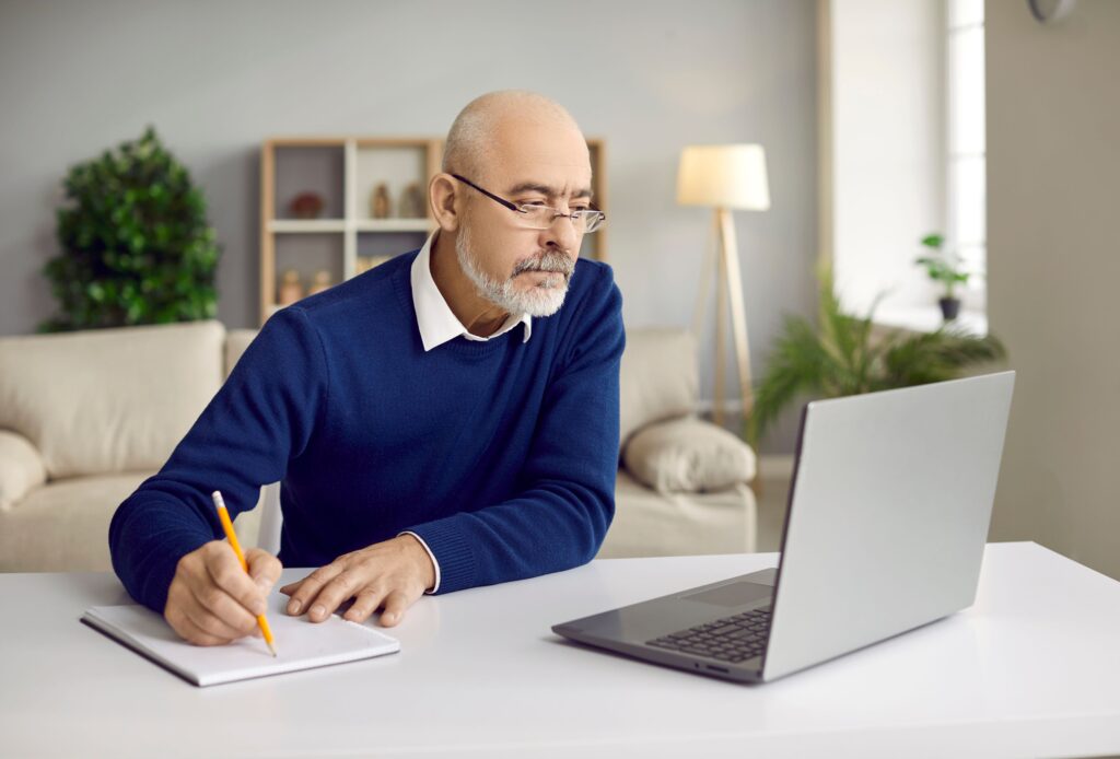 An older man does research on his laptop while taking notes.