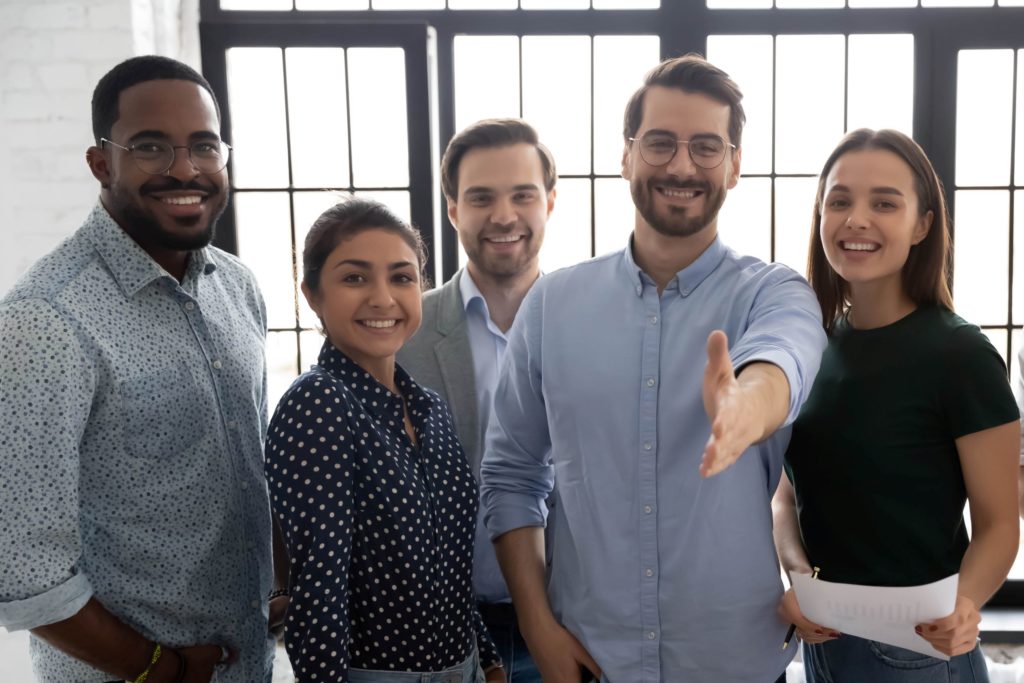 A diverse group of professionals stand together smiling while one extends their hand for a handshake.
