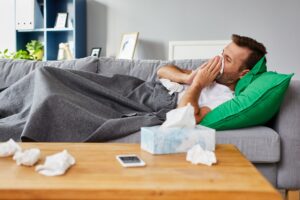 A sick man lays on his couch, blowing his nose and taking a sick day from work.