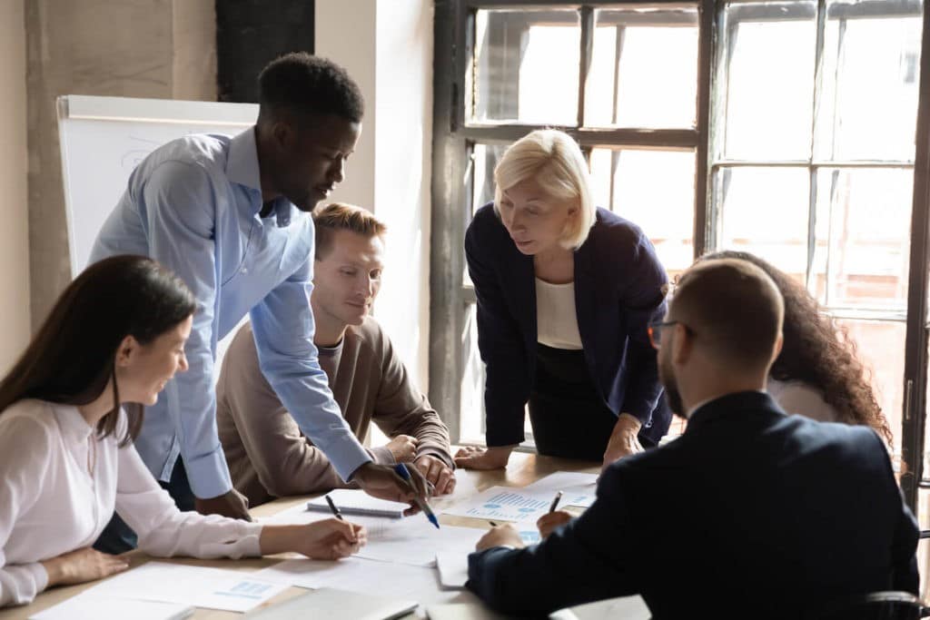 A diverse group of colleagues participate in a group meeting, going over paper work that is spread on the table between them.