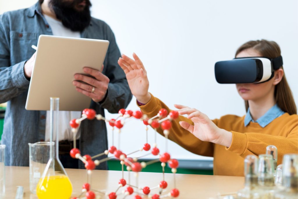 Girl using virtual reality headset in science class and teacher writing on tablet.