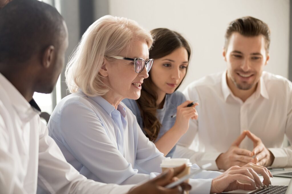 A group of people sit around a laptop while one speaks and points things out on the screen.