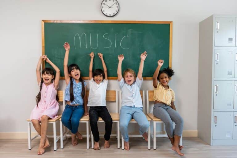 Student mental health: a group of students sit on a classroom table smiling and putting their hands in the air, the chalkboard has “music” written on it behind them.