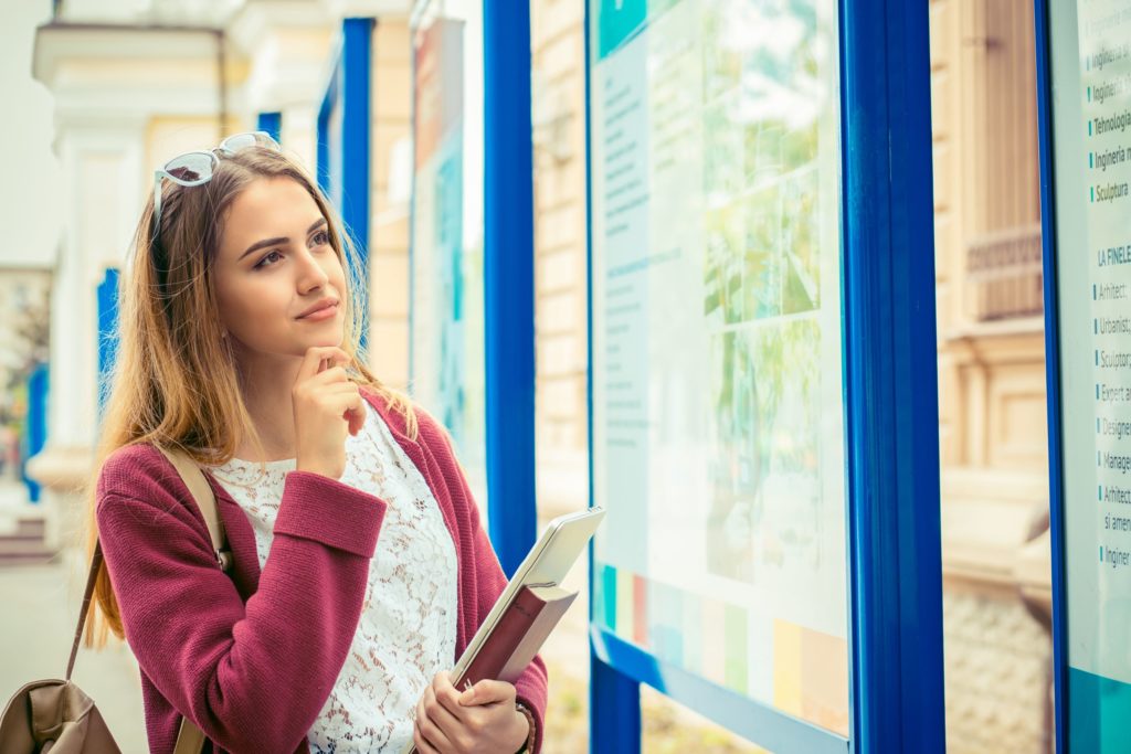 A future student stands with a book bag and books in her hands, looking at a informational college stand.