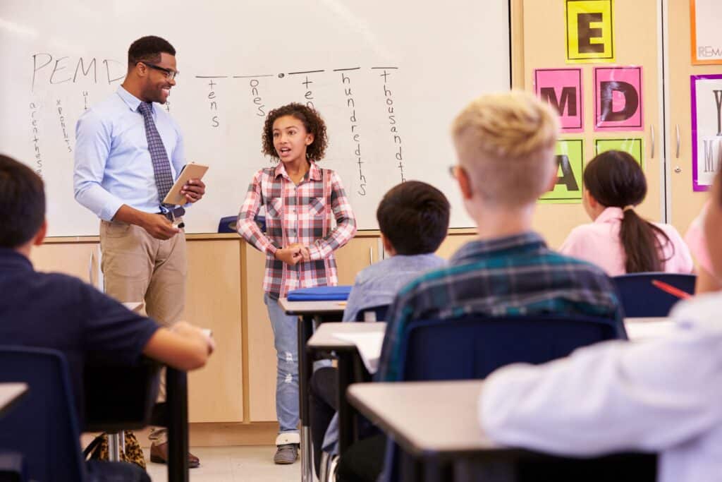 A student stands at the front of the classroom, doing a presentation while her teacher watches and smiles.