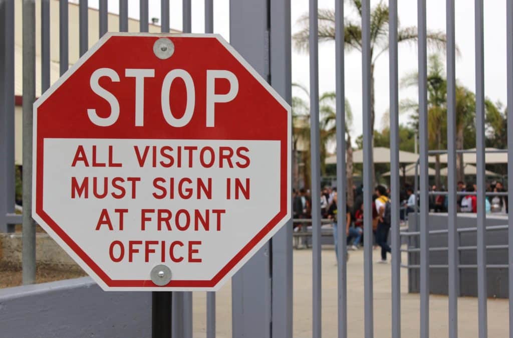 A sign hanging on a school gate reads, “Stop! All visitors must sign in at the office.”