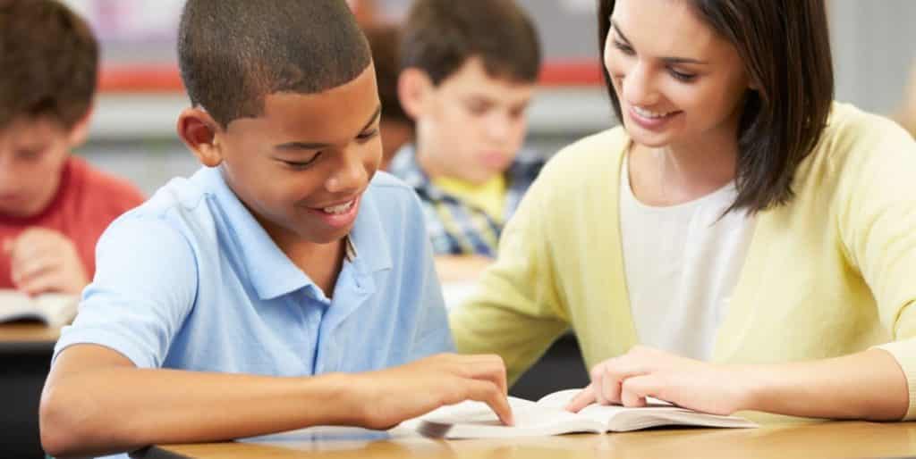 Teacher helping a young boy at his desk with a book.