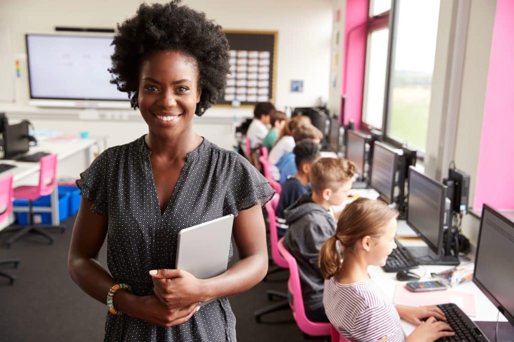Smiling teacher standing in a classroom filled with students using computers.