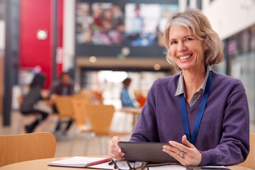 A teacher sits at a table, working on a tablet.