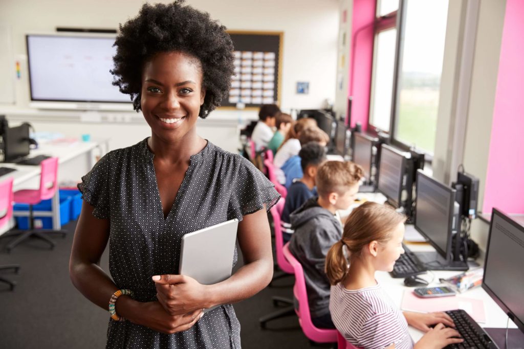 A female teacher holding a tablet teaches a line of high school students sitting at computers.