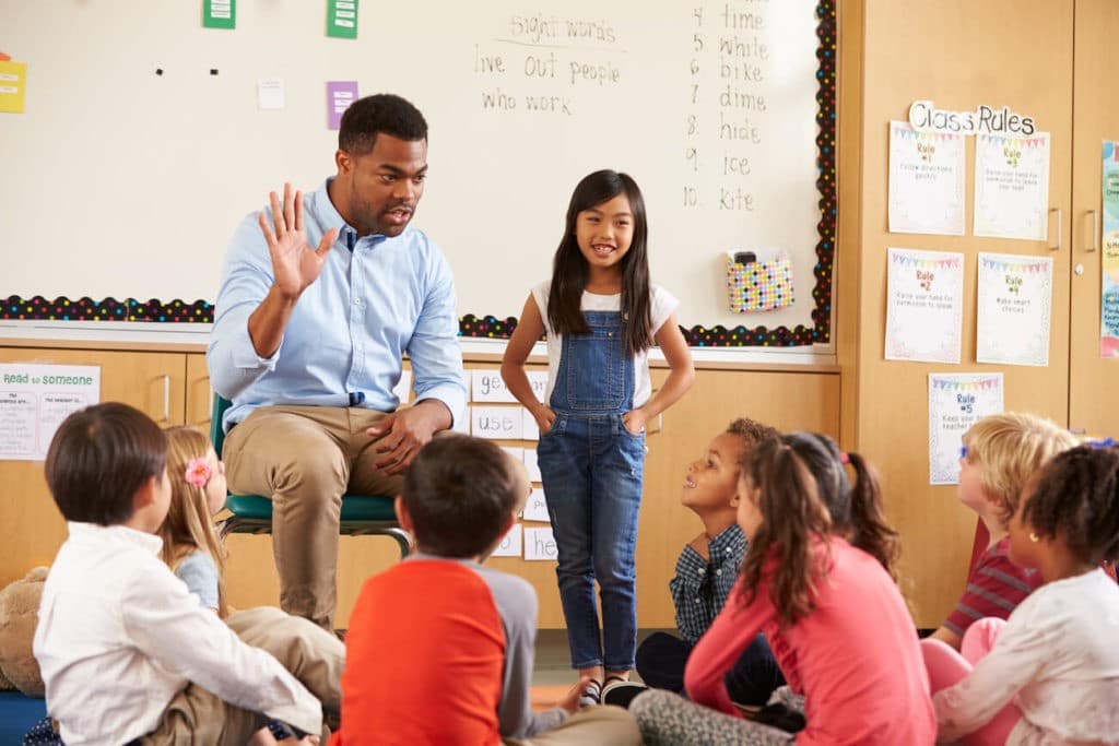 Teacher sitting with a group of students in a classroom asking a question.