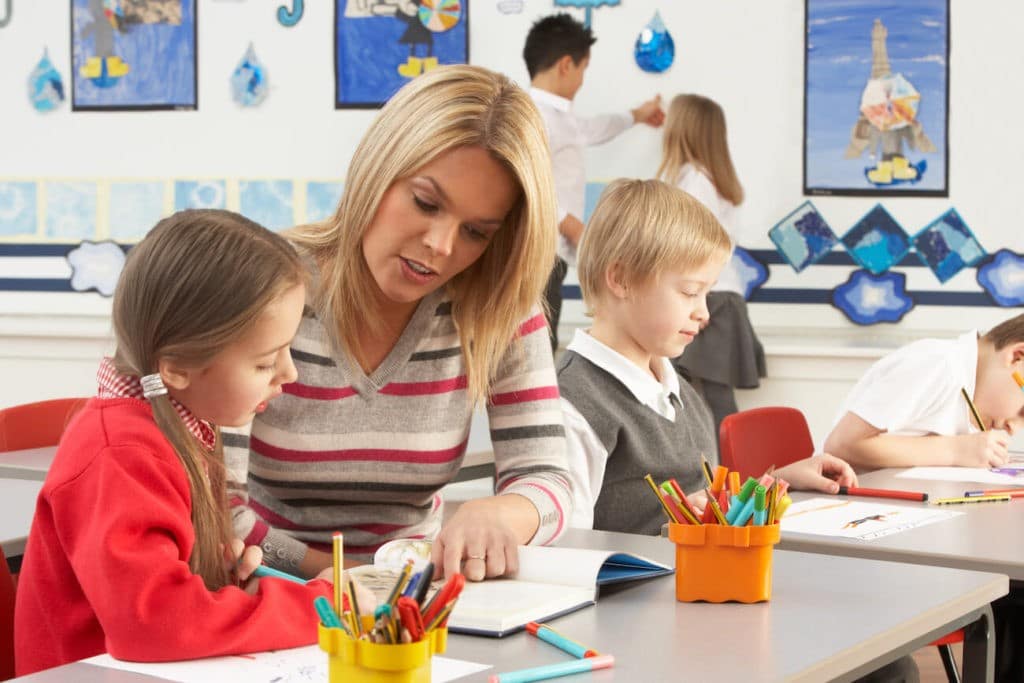 Teacher sitting at a desk with a student helping her read a book.